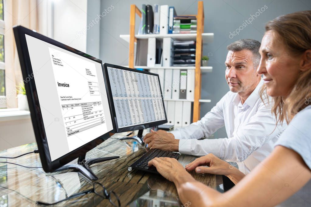 Side View Of Two Businesspeople Checking Invoice On Computer Over Desk At Workplace