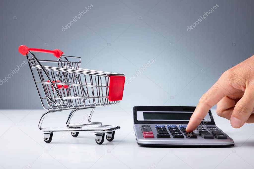 Close-up Of Empty Shopping Cart Near Person's Hand Using Calculator Over White Desk