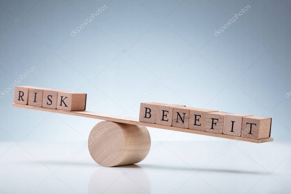 Close-up Of Risk And Benefit Wooden Blocks On Seesaw Against Reflective Desk