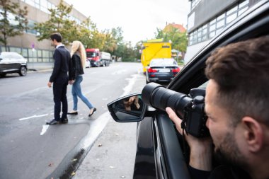 Genç bir erkek arabada oturmuş sokakta yürüyen bir çiftin fotoğrafını çekiyor.