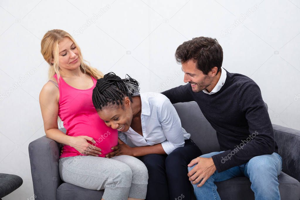 Smiling Young Man Looking At African Friend Listening To Pregnant Friend's Tummy Sitting On Sofa