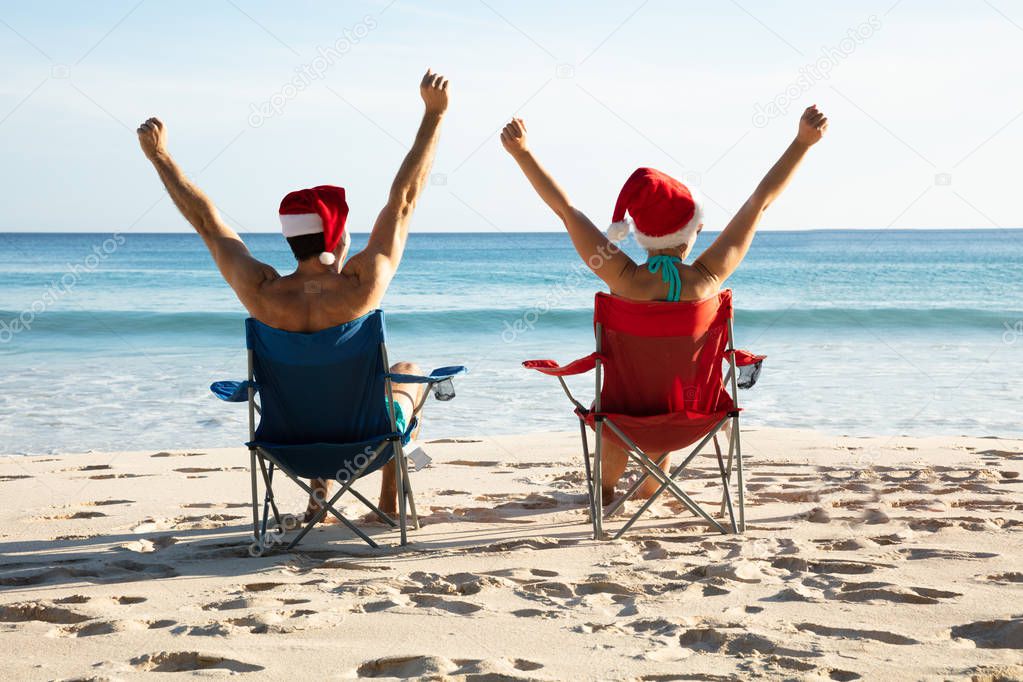 Rear View Of A Young Couple With Santa Hats Sitting On Chair Raising Their Arms