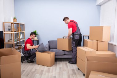Üniformalı iki genç nakliyeci karton kutulara ürün topluyor ve koyuyor.