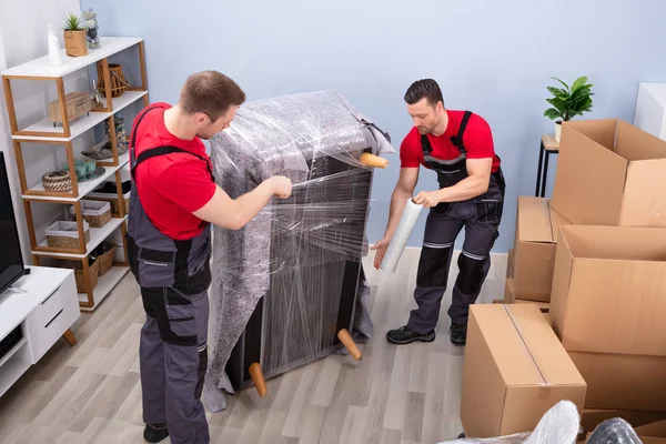 Deux Jeunes Déménageurs Masculins Enveloppant Canapé Avec Une Enveloppe Plastique — Photo