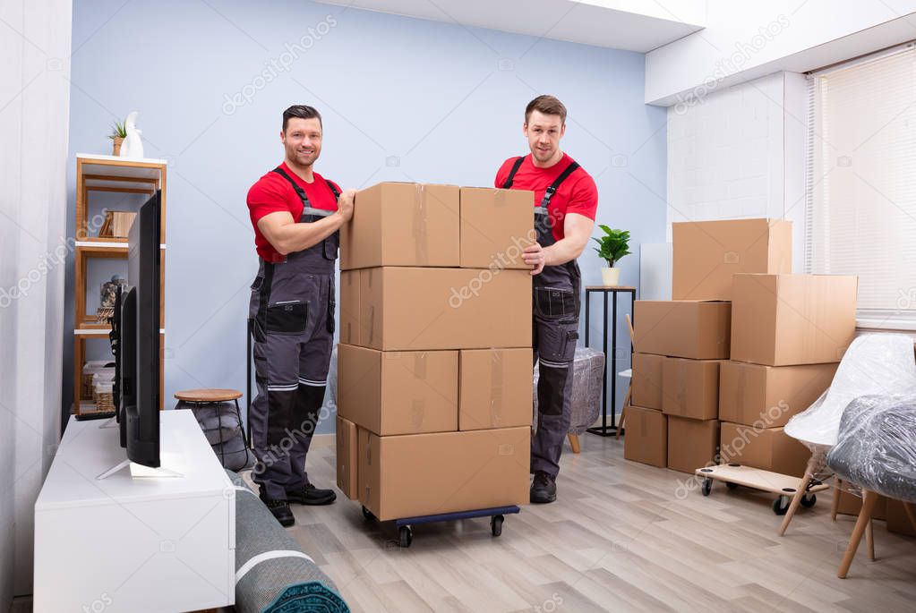 Smiling Professional Movers In Uniform With Stack Of Cardboard Boxes In The Living Room