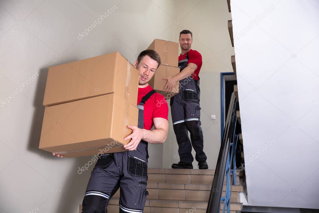 Portrait Of A Young Male Movers In Uniform Carrying Cardboard Boxes Walking Downward On Staircase