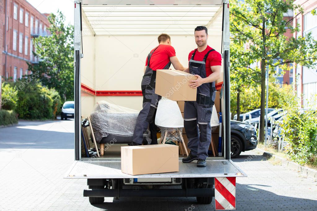 Two Young Men Unloading And Stacking The Brown Cardboard Boxes On Moving Truck