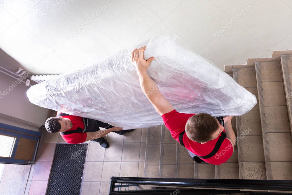 Two Young Male Movers In Uniform Carrying The Wrapped Mattress While Moving Downward The Staircase