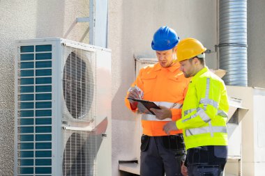 Güvenlik ceketi giyen iki elektrikçi çatıdaki havalandırmayı kontrol ediyor.