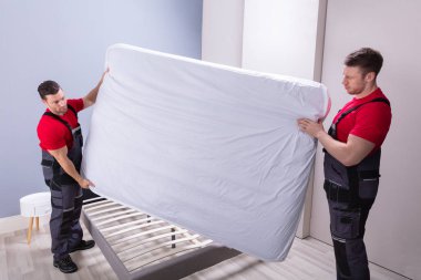 Üniformalı iki erkek profesyonel taşıyıcı yatağı yeni evdeki yatağın üzerine yerleştiriyor.