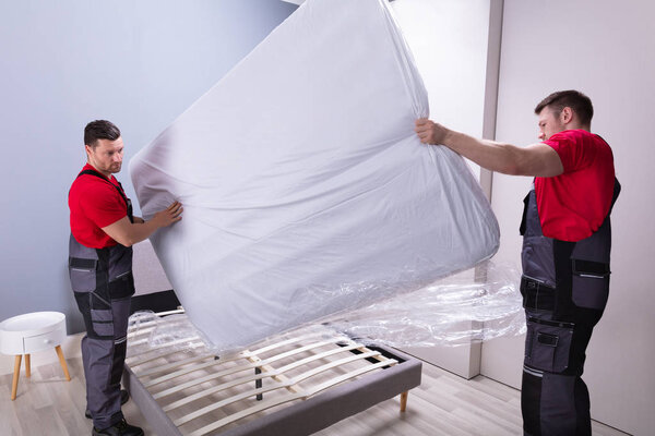 Two Male Professional Movers In Uniform Placing The Mattress Over The Bed In New House