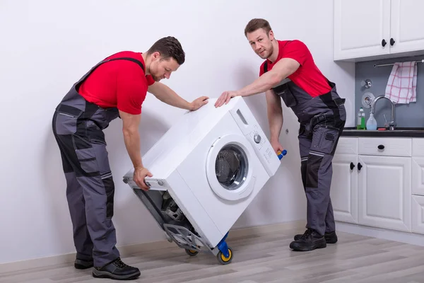 キッチンに現代の洗濯機を配置する制服の2人の若い男性の専門家の動き — ストック写真