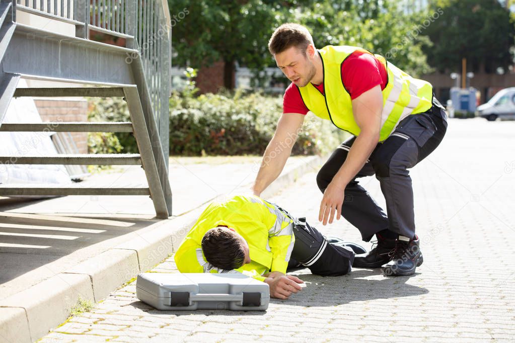 Worried Young Man Wearing Safety Jacket Looking At Person Falling On Street With Tool Box