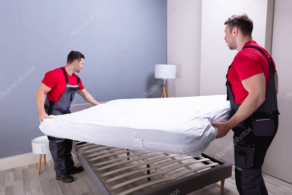 Two Male Professional Movers In Uniform Placing The Mattress Over The Bed In New House