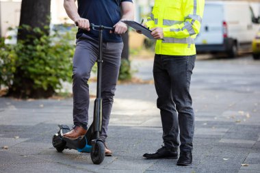 Memur elinde pano var. Şehrin caddesinde elektrikli scooter kullanan bir adamla duruyor.