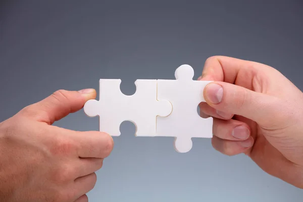 Close-up Of Two People\'s Hand Joining White The Jigsaw Puzzles Pieces On Gray Background