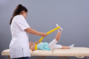 Klinikteki bir kadın fizyoterapistin verdiği sarı elastik bantla bacağını esneten kız.