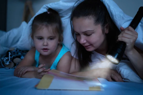 一位母亲把故事讲给躺在被子下面的女儿听 床头点亮了火把 — 图库照片