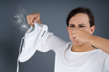Gri arka planda pis kokulu ayakkabılar tutan bir kadının burnunu kapattığı yakın plan.