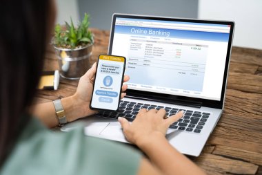 Dizüstü bilgisayardaki banka transferini doğrulamak için cep telefonu uygulaması kullanan kadın