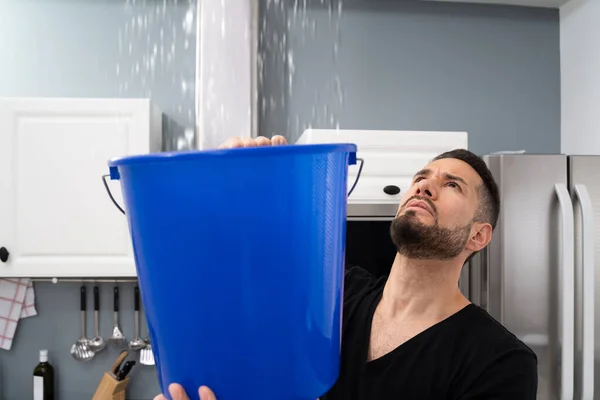 Беспокойный Человек Держит Ведро Пока Капли Воды Протекают Потолка Кухне — стоковое фото