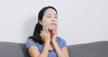 Genç kadın uygulamak yüz maskesi ve evde koltukta oturan