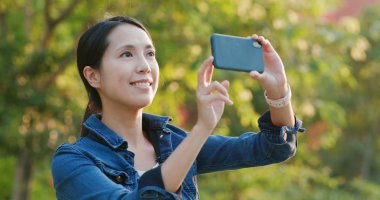 Güneş ışığı ve flare altında yeşil arka plana sahip fotoğraf almak için cep telefonu kullanan kadın