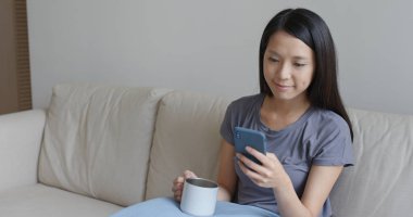 Kadın cep telefonu kullanıyor ve evdeki kanepeye oturuyor.