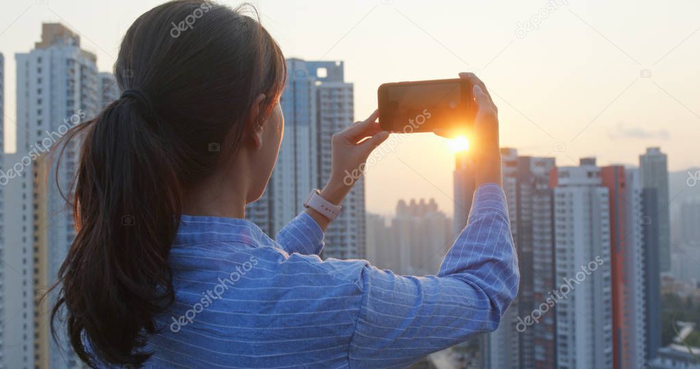 Woman enjoy the view of the city and take picture on mobile phone under sunset view