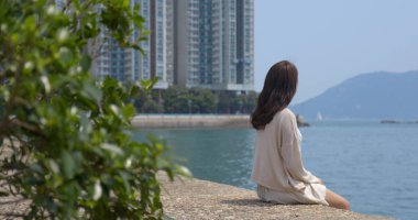 Kadın deniz kenarında oturup şehir manzarasının ve denizin tadını çıkarıyor.