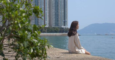 Kadın deniz kenarında oturup şehir manzarasının ve denizin tadını çıkarıyor.
