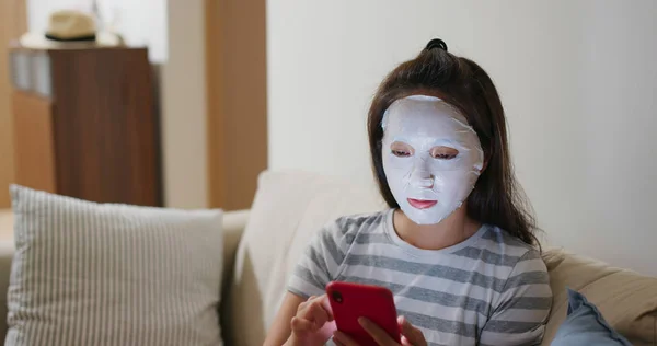 Vrouw toepassen gezichtsmasker en gebruik van mobiele telefoon thuis — Stockfoto