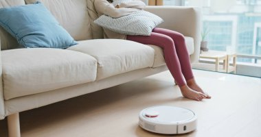 Kadın ayaklarını kaldırıp evdeki yere robot süpürgeyle vuruyor.
