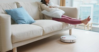 Kadın ayaklarını kaldırıp evdeki yere robot süpürgeyle vuruyor.