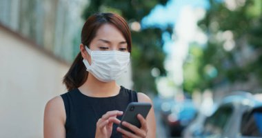 Kadın yüz maskesi takıyor ve şehirde cep telefonu kullanıyor.