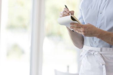 Genç garson restoranda dururken not defteri yazma midsection