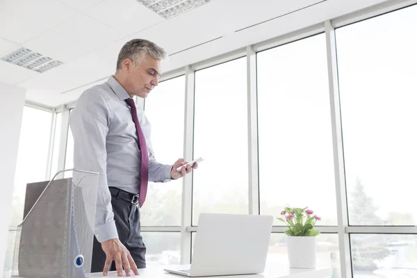 新しいオフィスの机に立ちながらスマートフォンを使う成熟したビジネスマン — ストック写真