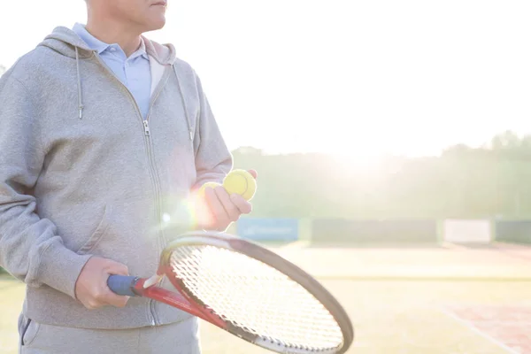 Midsection of mature man holding tennis balls and racket on court against clear sky