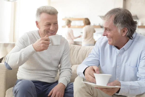Uśmiechnięci Dojrzali Mężczyźni Trzymający Kawę Siedząc Kanapie Domu — Zdjęcie stockowe
