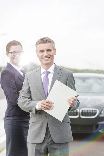 クリップボードと実業家故障車に対して立っている笑顔の保険代理店の肖像画 — ストック写真
