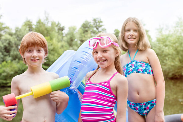 Portrait of smiling friends in swimwear standing with pool raft and squirt gun at lakeshore