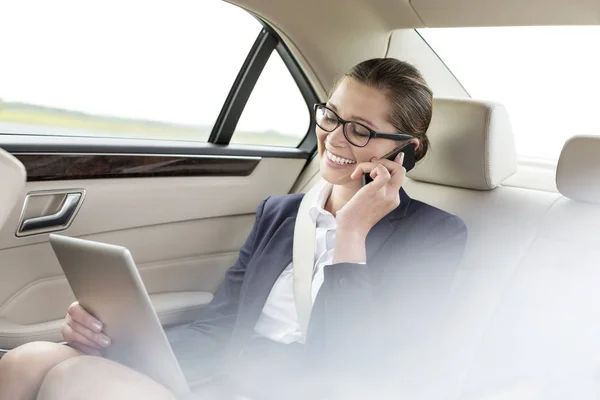 微笑的女商人在汽车中使用数字平板电脑时在智能手机上说话 — 图库照片