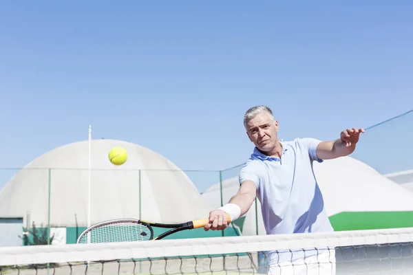 澄んだ青い空に対して裁判所にラケットでテニスボールを打つ自信がある中年の男性 — ストック写真