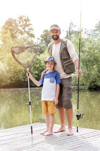 Full length portrait of smiling father and son standing with fishing tackles on pier against lake
