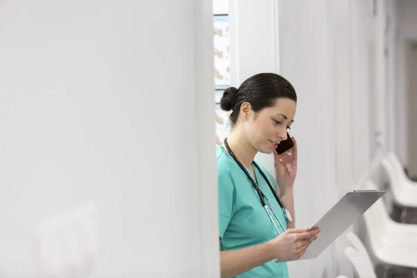 Nurse talking on smartphone while reading document on clipboard at hospital