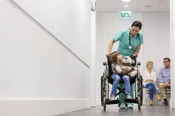 病院の廊下で待っている患者の間に車椅子にテディベアを持つ看護師が少年を押す — ストック写真
