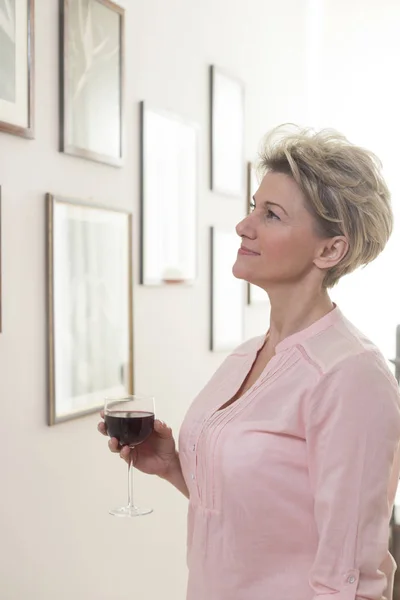 Mature Woman Holding Wineglass While Looking Picture Frames Wall — Stock Photo, Image