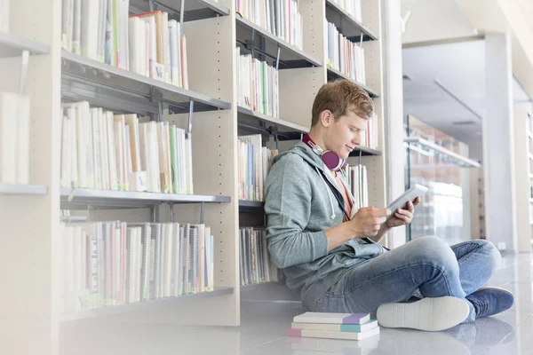 学生坐在图书馆的地板上 用数字平板电脑对书架 — 图库照片