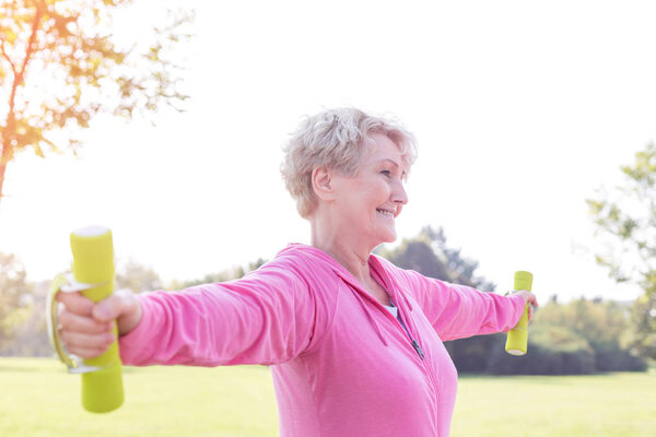 Senior woman with arms outstretched lifting dumbbells in park