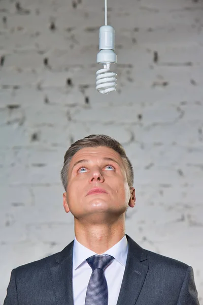 オフィスのレンガの壁に対してぶら下がっエネルギー効率の良い電球を見て思慮深い成熟したビジネスマン — ストック写真